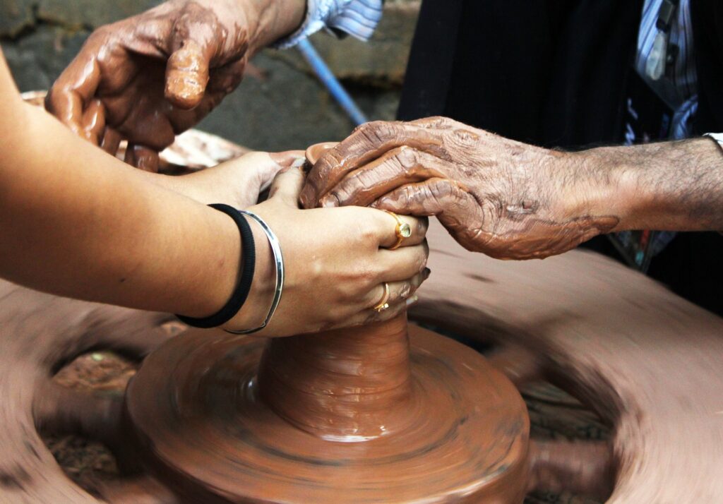 The hands of two individuals work together to form a piece of pottery on a pottery wheel.